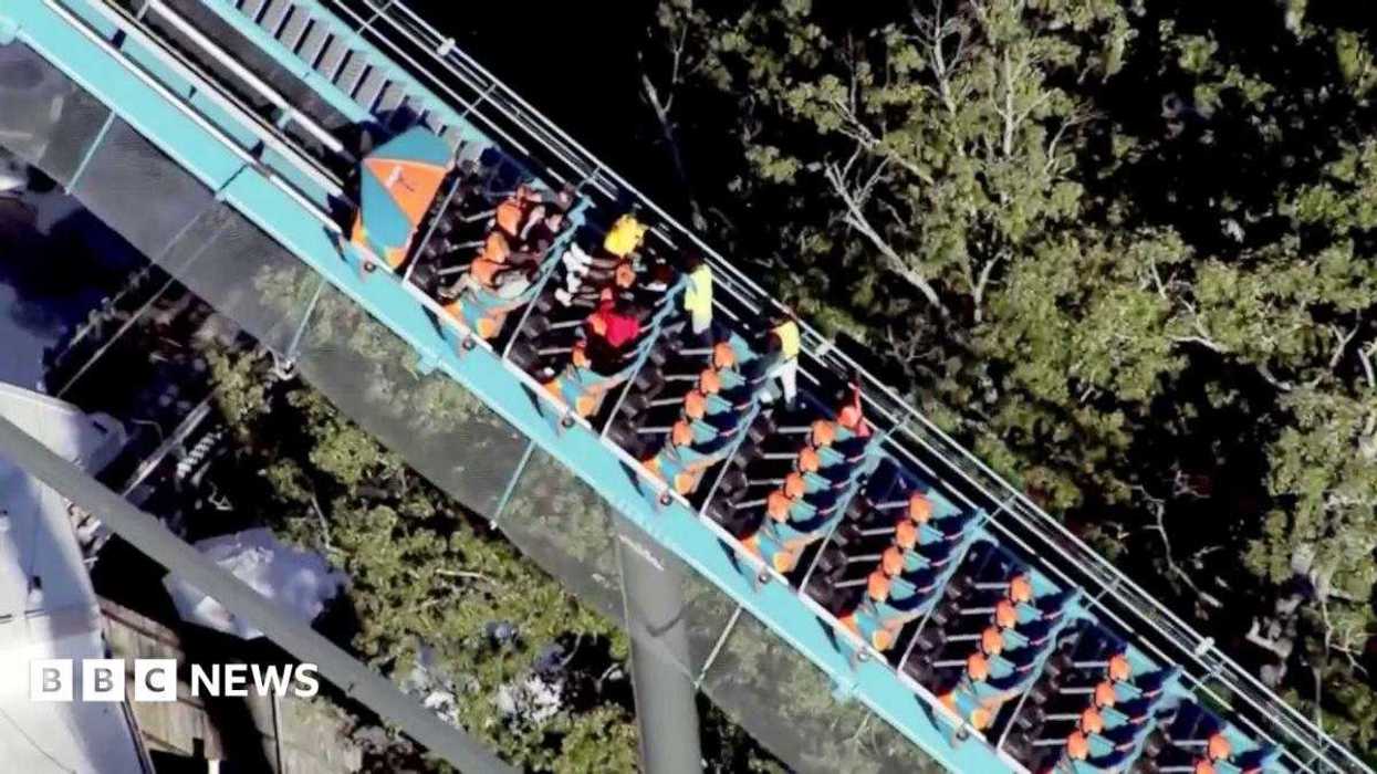 Vista aérea de una montaña rusa de riel azul verdoso con un tren de pasajeros y dos personas con chalecos amarillos