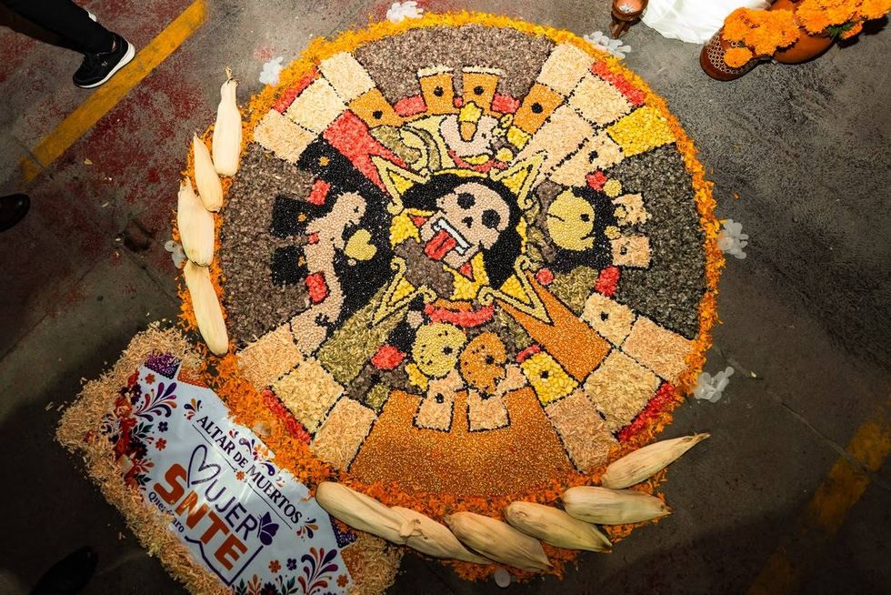 Vista cenital de un tapete de semillas y granos rodeado de flor de cempas\u00fachil, con el letrero \u201cAltar de Muertos Mujer SNTE Quer\u00e9taro\u201d.