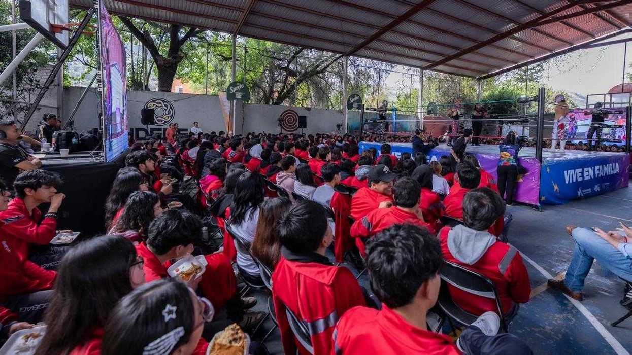 Vista general de estudiantes sentados frente a un ring de lucha libre, con un templete que dice \u201cVive en familia\u201d