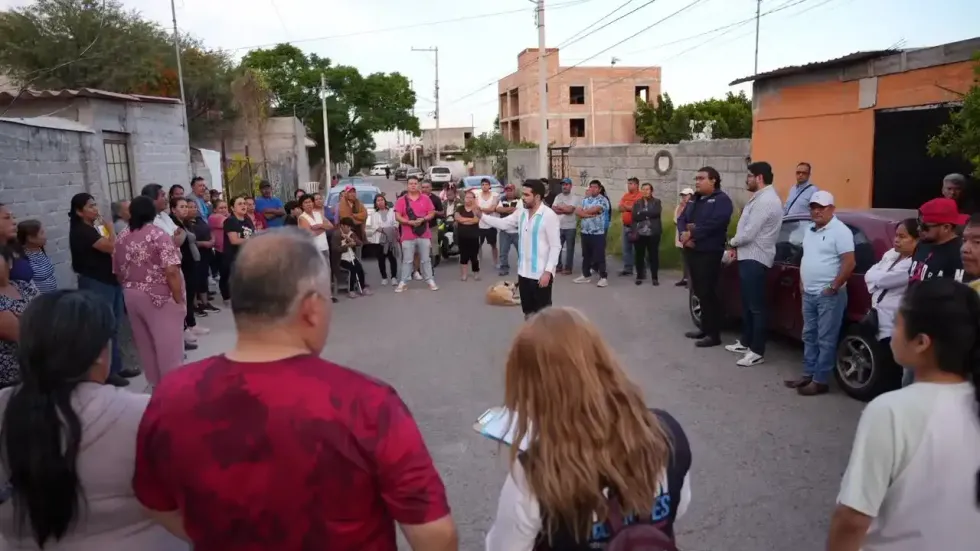 Vista general de la asamblea vecinal en el asentamiento Los Pinos: decenas de residentes forman un c\u00edrculo en una calle sin pavimentar mientras el senador Dorantes se dirige a ellos.
