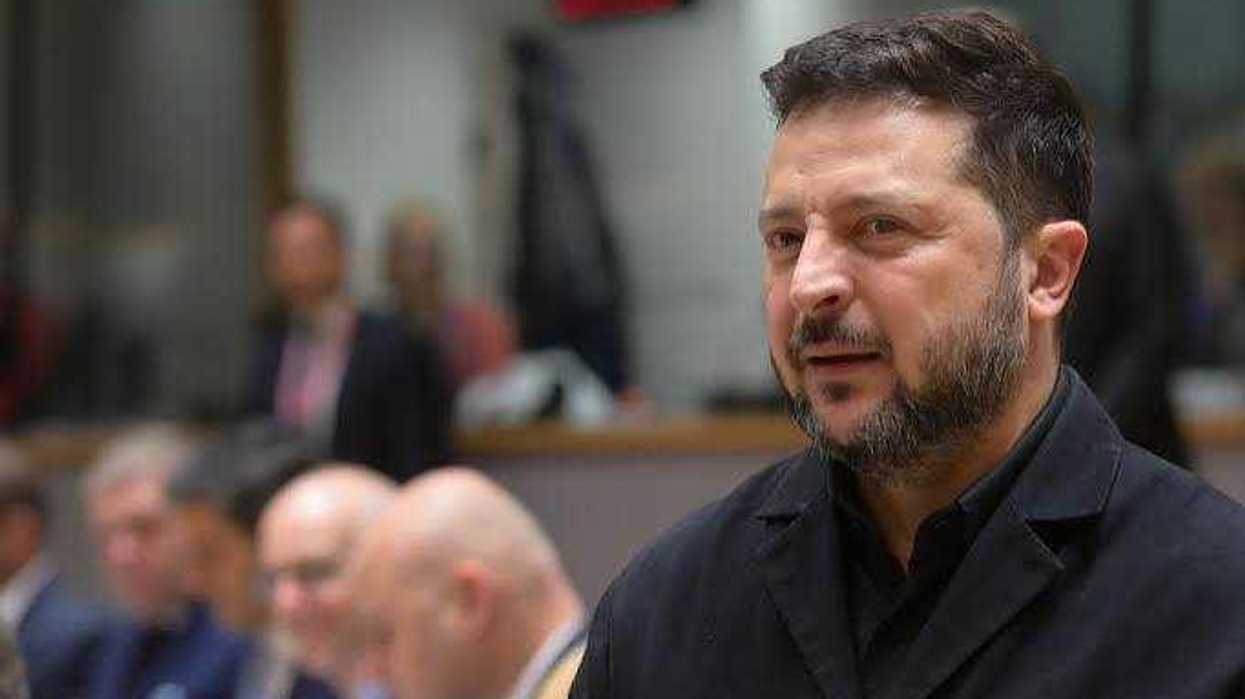 Volodímir Zelenski, presidente de Ucrania, de barba y chamarra oscura, en un salón con personas desenfocadas al fondo
