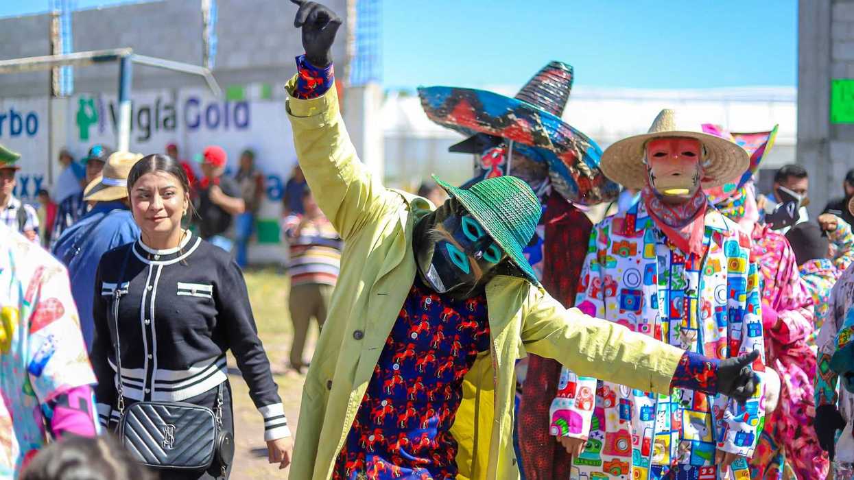 Xhitases con máscaras y atuendos de colores avanzan entre el público al aire libre; uno, con gabardina amarilla y sombrero verde, levanta el brazo.